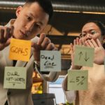 Two professionals organize tasks on glass using sticky notes in an office setting.