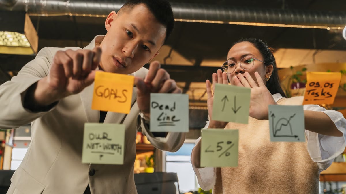 Two professionals organize tasks on glass using sticky notes in an office setting.